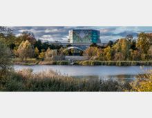 Ein moderner Glasbau mit geometrischer Fassade erhebt sich hinter einem bewaldeten Park mit herbstlichem Laub, einem kleinen Pavillon und einem See im Vordergrund, umgeben von Schilf.