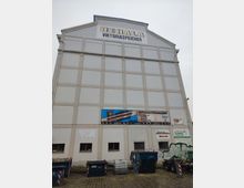 Ein großes, weißes Lagergebäude mit der Aufschrift „BEHALA Viktoriaspeicher“ auf der obersten Ebene. Die Fassade ist schlicht mit mehreren kleinen Fenstern im unteren Bereich, und vor dem Gebäude stehen Müllcontainer und Recyclingbehälter auf einem gepflasterten Hof.