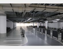 Unterirdische Fahrradparkgarage mit markierten Fahrspuren, abgestellten Fahrrädern und metallischen Absperrungen. Die Decke zeigt sichtbare Lüftungsrohre und Beleuchtungskörper.