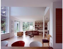 Ein Wohnzimmer mit modernen Möbeln, großen Fensterfronten und Blick auf einen grünen Garten. Der Raum verfügt über eine Bücherwand, Holzboden, gemütliche Sofas sowie eine offene und helle Gestaltung.