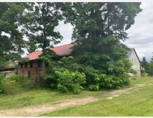 Backsteingebäude mit roten Dachziegeln, umgeben von großen Bäumen und dichtem Grün. Im Vordergrund ein unbefestigter Weg und eine Wiese; rechts neben dem Hauptgebäude ein weiteres Haus mit weißer Fassade.