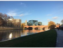 Flusslandschaft mit modernem und historischem Hintergrund: Ein breiter Fluss mit reflektierendem Wasser und einer roten Backsteinbrücke mit Bögen. Im Hintergrund sind moderne Glasgebäude, ein DB-Logo und weinende Weiden entlang des Ufers zu sehen. Ein Gehweg läuft parallel zum Fluss.