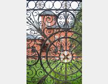 Ein kunstvoll verziertes Schmiedeeisentor mit floralen und spiralförmigen Mustern, durch das unscharf ein rotes Backsteingebäude mit runden Fenstern und einem spitzen Dach sichtbar ist. Im Vordergrund ist grüne Rasenfläche zu erkennen.