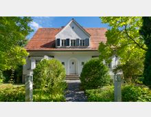 Einfamilienhaus mit weißer Fassade, rotem Satteldach und blauen Fensterläden, umgeben von einem gepflegten Garten mit dichtem Grün. Ein gepflasterter Weg führt durch den Vorgarten zu einer weißen Eingangstür mit Rundbogen.