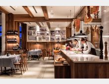 Ein gemütliches Restaurant mit Holzelementen, karierten Tischdecken und Regalen voller Weinflaschen. Im Vordergrund eine Theke mit Brot und Zutaten, dahinter zwei Köche beim Zubereiten von Speisen; an der Decke hängen Schinken und getrocknete Kräuter.