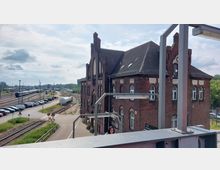 Backsteingebäude eines Bahnhofs mit historischen Details, daneben Bahngleise mit abgestellten Zügen und ein Parkplatz. Im Vordergrund eine erhöhte Plattform mit Geländern und teilweise verdeckter Sicht auf das Gebäude.