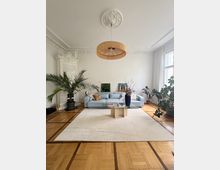 Helles Wohnzimmer mit hohen Decken, Stuckverzierungen und großem Deckenrosetten-Leuchtenelement, mit Parkettboden und großem hellen Teppich. Vor einer hellblauen Sofa-Sitzgruppe steht ein niedriger Glastisch; große Zimmerpflanzen und bodentiefe Fenster mit weißen Vorhängen rahmen den Raum.