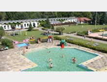 Ein Außenbereich eines Ferienparks mit einem kleinen rechteckigen Schwimmbecken im Vordergrund, in dem mehrere Menschen baden. Im Hintergrund sieht man eine große Rasenfläche mit Sitzmöglichkeiten, Spielgeräten und Gebäuden mit wellenförmigen Dachkanten, die von Bäumen umgeben sind.