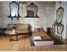 Alt-Text: Ein Raum mit abgenutzten, beigen Wänden und Holzboden, eingerichtet mit einem antiken Sofa, einem kleinen Holztisch mit einer Vase voller Rosen, einer Matratze mit Decke und Kissen sowie einem goldenen Schminktisch mit Spiegel. Zwei verzierte Wandspiegel hängen über dem Sofa.