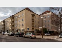 Wohnblock mit vier Stockwerken in heller Putzfassade und roten Ziegeldächern, mit Balkonen und begrünten Eingangsbereichen. Im Vordergrund eine ruhige Straßenansicht mit parkenden Autos und Laternen, daneben Gehwege und spärliche Vegetation.