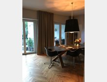 Esszimmer mit einem dunklen rechteckigen Tisch und modernen schwarzen Stühlen auf einem hölzernen Fischgrät-Parkettboden. Große Fenster mit bodenlangen beigefarbenen Vorhängen führen zu einer Terrasse mit Pflanzen, und eine große schwarze Pendelleuchte hängt über dem Tisch.