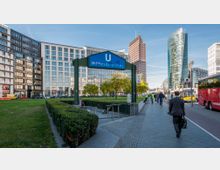 Eingang zur U-Bahn-Station "Bahnhof Potsdamer Platz" in Berlin, umgeben von modernen Bürogebäuden und Grünflächen; rechts im Bild verläuft ein Gehweg mit Fußgängern, im Hintergrund sind markante Hochhäuser und ein roter Reisebus zu sehen.