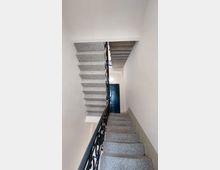 Treppenhaus mit grauen Steinstufen und einem schwarzen Geländer aus geschwungenem Metall. Die Wände sind weiß, und im oberen Bereich ist eine geschlossene dunkle Tür sichtbar.