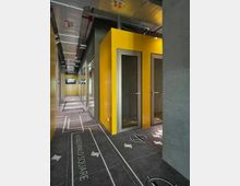 Schmaler Flur in einem modernen Gebäude mit gelben Kabinenwänden und grauem Bodenbelag, der mit Grafiken und Beschriftungen wie „Königswald Square“ verziert ist. An der Decke befinden sich freiliegende Lüftungsrohre und Kabelkanäle.