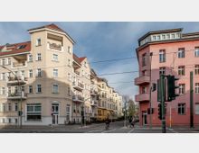 Straßenecke mit mehrstöckigen Wohnhäusern im typischen Altbaustil: links ein beiges Gebäude mit Balkonen, rechts ein rosafarbenes Haus. Im Vordergrund eine Kreuzung mit Ampeln und Verkehrsschildern, im Hintergrund Bäume und blauer Himmel.