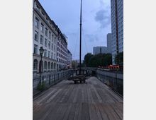 Holzplattform am Ufer eines schmalen Kanals bei Dämmerung, flankiert von mehrstöckigen Gebäuden auf beiden Seiten. Im Hintergrund ist eine Brücke über den Kanal und eine spärlich beleuchtete Straße mit Laternen und Geschäften zu sehen.