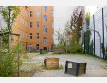 Hinterhof einer Wohnanlage mit gepflastertem Boden, kleinen Grünflächen, Bäumen und Sitzbänken. Im Hintergrund ein orangefarbenes Gebäude mit großen Fenstern sowie eine helle Hauswand mit Kletterpflanzen und einem Basketballkorb.