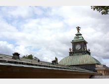 Gebäudedetail mit einer kupfergedeckten Kuppel und einem Uhrenturm, der von einer Adlerfigur und Statuen verziert ist, vor einem bewölkten Himmel. Im Vordergrund ist ein geneigtes Dach mit mehreren Schornsteinen zu sehen.