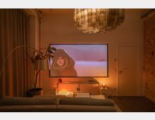 Wohnzimmer mit gemütlicher Beleuchtung, einem Projektorbildschirm an der Wand, der eine Filmszene zeigt, und moderner Einrichtung, darunter ein Sofa, ein kleiner Tisch, eine Stehleuchte, und ein Regal mit Dekorationen und Elektronik. Links ist ein Vorhang und rechts eine Tür zu sehen, die Wand weist teilweise unverputztes Mauerwerk auf.