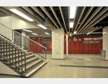 U-Bahnhof Berliner Straße mit rot verkleideten Wänden, einem Treppenaufgang links, einer Übersichtskarte der Linie U7 an einer wandmontierten Tafel und einem beleuchteten Deckenraster mit Leuchtstofflampen.