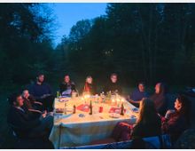 Eine Gruppe von Menschen sitzt bei Dämmerung um einen Tisch im Freien, der mit einer Tischdecke gedeckt ist und von mehreren Kerzen beleuchtet wird. Im Hintergrund sind dichte Bäume und ein dunkler Himmel sichtbar.
