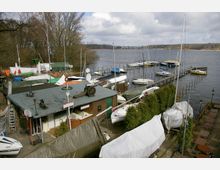 Kleiner Jachthafen an einem See mit zahlreichen Segelbooten, die teils im Wasser und teils auf Land abgedeckt gelagert sind. Im Vordergrund befindet sich ein Flachdachgebäude umgeben von festgemachten Booten, Planen und Pflanzen; im Hintergrund erstreckt sich der See mit einem Steg und weiteren Booten.