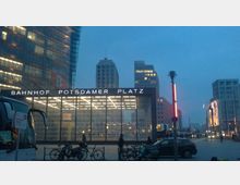 Außenansicht vom Bahnhof Potsdamer Platz in Berlin bei Dämmerung, mit beleuchtetem Glasdach und umgeben von modernen Hochhäusern; im Vordergrund parken Fahrräder und Autos.