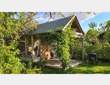 Kleiner Holzpavillon mit großem Fenster und Satteldach, umgeben von dichtem Grün, Bäumen und Sträuchern. Im Vordergrund stehen Gartenmöbel auf einer Terrasse und ein Grill auf einem Rasenstück.