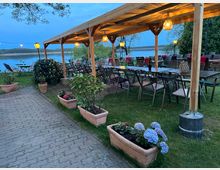 Eine überdachte Terrasse mit Holzkonstruktion und Hängelampen, die mehrere Tische und Stühle umfasst, steht auf einer Rasenfläche nahe einem ruhigen See. Blumenkübel mit verschiedenen Pflanzen säumen den gepflasterten Weg, und im Hintergrund sind Bäume und eine blaue Abenddämmerung sichtbar.