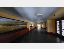 Langer, schmaler Innenraum mit gelben Wänden, roten Holztheken und einer Garderobenanlage links. Auf der rechten Seite befinden sich Glastüren und ein Heizkörper; der Raum wird von rechteckigen Deckenleuchten beleuchtet.