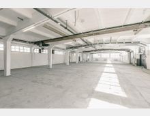 Weitläufige, helle Industriehalle mit weißen Wänden und mehreren Fensterreihen, die Tageslicht hereinlassen. Sichtbare Stahlträger, eine Kranbahn mit einer Traglast von 2000 kg und ein Betonboden prägen den Raum.