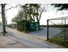 Gepflasterter Gehweg an einer Straße mit Bäumen, neben einem Metallzaun, der ein Gelände mit einem kleinen grünen Häuschen und einer Einfahrt umgibt. Im Hintergrund sind Laternenmasten und Hecken sichtbar.