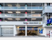 Mehrstöckiges Parkhaus mit der Beschriftung „Q-Park Forum Steglitz“ über dem Haupteingang. Der Eingang führt in eine Tiefgarage, daneben befinden sich Fahrradständer und Beschilderungen, die freie Parkplätze und Einfahrtshinweise anzeigen.