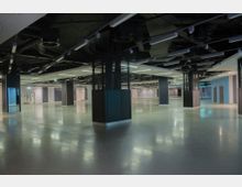 Weitläufiger, leerer Innenraum mit glänzendem Boden, zahlreichen quadratischen Säulen und einer offenen Decke, die Rohrleitungen und Beleuchtungskörper sichtbar zeigt. Der Raum ist von neutralen Farben, künstlichem Licht und modernen architektonischen Elementen geprägt.