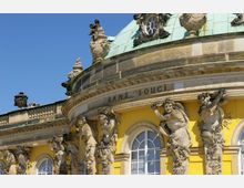 Detailaufnahme des Schlosses Sanssouci in Potsdam, Deutschland, mit gelber Fassade, weißen Rundbogenfenstern, kunstvollen Steinskulpturen und einem kupfergrünen Kuppeldach. Über den Skulpturen ist der Schriftzug „Sans, Souci“ in die Fassade eingraviert.
