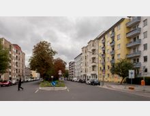 Straßenszene in einem Wohnviertel mit mehrstöckigen Wohnhäusern auf beiden Seiten, rechts gelbe Fassaden mit Balkonen. Eine Verkehrsinsel mit Bäumen und Schildern sowie parkende Autos säumen die Straße, die eine 30-km/h-Geschwindigkeitsbegrenzung aufweist.