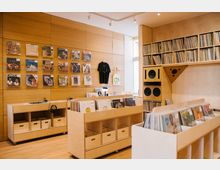 Ein Plattenladen mit hellen Holzmöbeln, offenen Regalen voller Schallplatten und einer Wand mit Schallplattencovern auf einer perforierten Holzverkleidung. Im Hintergrund befinden sich Lautsprecher und Regale mit weiteren Schallplatten; eine große Fensterfront sorgt für natürliche Beleuchtung.