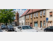 Straßenszene in einer Kleinstadt mit geparkten Autos, historischen Gebäuden mit roten Ziegeldächern und einem Rundturm aus Backstein im Hintergrund. Ein Baum spendet Schatten, und ein Verkehrsschild weist auf Parkzeiten hin.