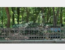 Ein metallisches, dekoratives Geländer mit spiralförmigen Verzierungen steht vor einem dichten, grünen Park mit hohen Bäumen und Sträuchern. Im Hintergrund sind schattige Waldflächen und ein blauer Gegenstand zu erkennen.