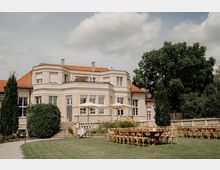 Helles, zweigeschossiges Herrenhaus mit rotem Ziegeldach, großen Sprossenfenstern und Balkonterrasse, davor eine erhöhte Steinterrasse mit Sonnenschirmen. Im Vordergrund liegt ein gepflegter Rasen mit aufgestellten Stuhlreihen und Tischen, umgeben von Büschen und großen Bäumen.