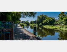 Gepflasterter Uferweg entlang eines ruhigen Flusses, umgeben von dichtem Grün und Bäumen, mit Graffiti auf einer Ziegelmauer links. Im Hintergrund sind vereinzelte Gebäude und ein Strommast sichtbar.