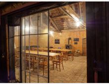 Blick durch ein großes Fenster mit Metallrahmen in einen rustikalen Raum mit Backsteinwänden, Holzbalkendecke und Steinboden. Im Raum stehen Holztische und Stühle, ein Holzofen und gestapeltes Brennholz sind sichtbar.