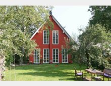 Ein rotes Einfamilienhaus mit Satteldach, großen bodentiefen Fenstern und einer gepflegten Rasenfläche im Vordergrund. Der Garten ist von Bäumen umgeben, mit einer Schaukel links und einem Gartentisch mit Stühlen rechts.