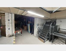 Ein Lagerraum mit unverputzten Ziegelwänden, Regalen und verschiedenen gestapelten Gegenständen wie Holz- und Metallteilen. Eine helle Deckenleuchte beleuchtet den Raum, der auch eine abgesenkte Decke und sichtbare Kabel zeigt.