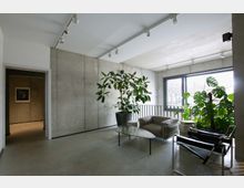 Moderner Innenraum mit Sichtbetonwänden, großen Fenstern und minimalistischer Einrichtung. Der Raum enthält Pflanzen, Sessel, einen ovalen Couchtisch und eine schwarze Metallstuhlgruppe vor einem Geländer.