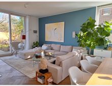 Helles Wohnzimmer mit einer großen beige-farbenen Eckcouch, blauer Akzentwand mit abstraktem Gemälde, und einer Zimmerpflanze in einem weißen Topf. Links befindet sich eine Glasfront mit Blick auf den Garten, während ein Esstisch mit weißen Stühlen im Vordergrund steht.