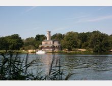 Historische Kirchengebäude mit einem quadratischen Turm, umgeben von Wald und gelegen an einem Flussufer; im Vordergrund fließt das Wasser, mit einem Boot nahe der Kirche.