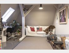 Gemütlicher Wohn- und Arbeitsbereich in einem Dachgeschosszimmer mit sichtbaren Dachbalken. Links steht ein Schreibtisch mit einem Stuhl und Pflanzen, rechts eine weiße Couch mit Kissen im US-Flaggen-Design, daneben ein Indoor-Rudergerät und ein Teppich, der den Boden bedeckt.