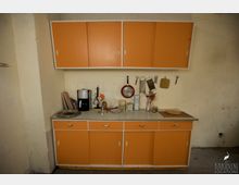 Küchenzeile in einem schlichten Raum mit orangefarbenen Schranktüren oben und unten, einer Arbeitsplatte mit Küchenutensilien wie einer Kaffeemaschine und Brettchen sowie an der Wand hängenden Kochutensilien wie Sieben und Pfannen. Der Putz der Wände ist teilweise abgenutzt.