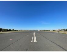 Ehemalige Start- und Landebahn eines Flughafens, umgeben von einer weitläufigen, offenen Grünfläche mit vereinzelten Bäumen und Gebäuden im Hintergrund unter einem klaren blauen Himmel.